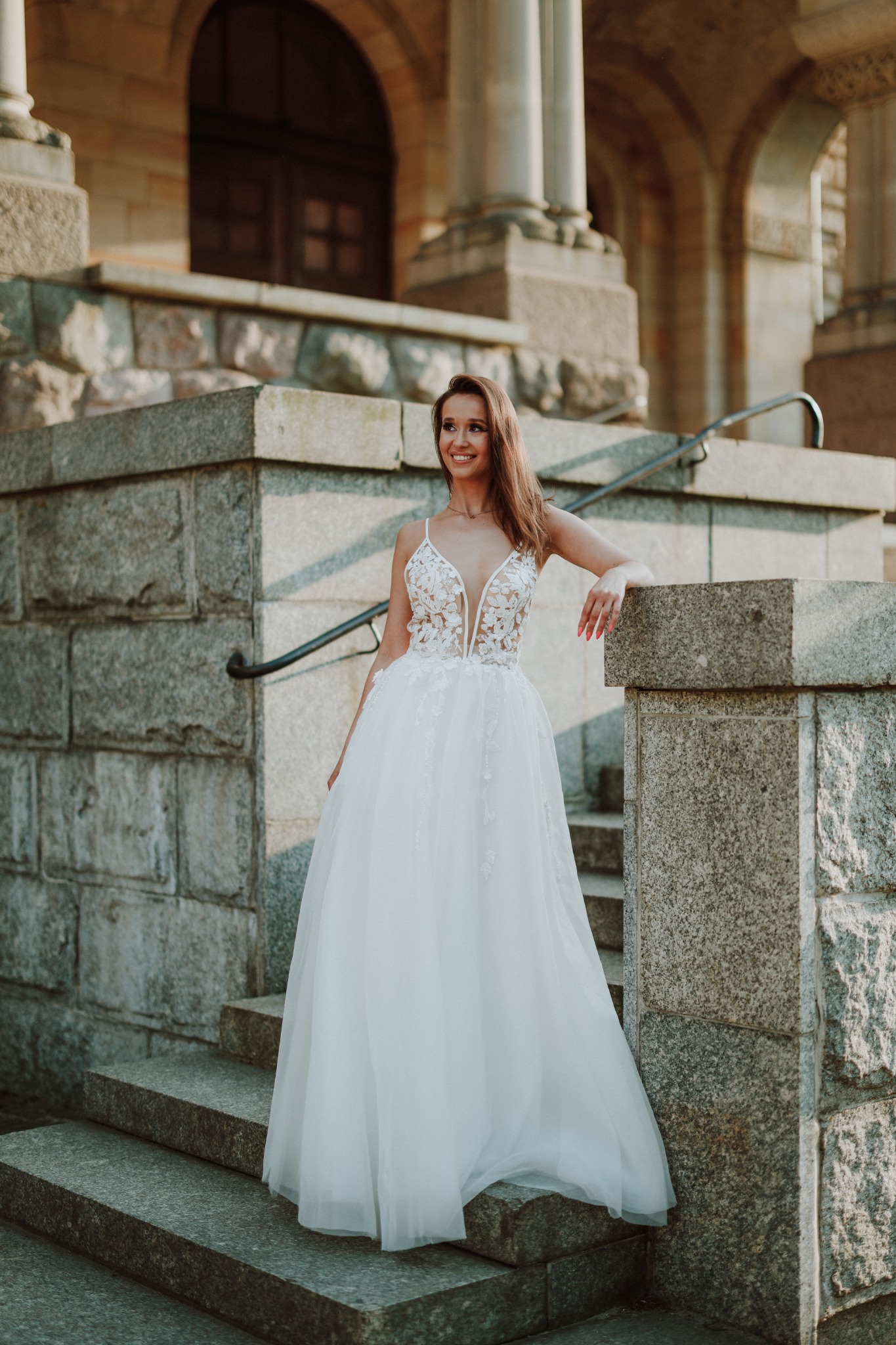 Uśmiechnięta kobieta w białej, eleganckiej sukni z koronkową górą, stojąca na kamiennych schodach przed budynkiem z kolumnami i łukami.