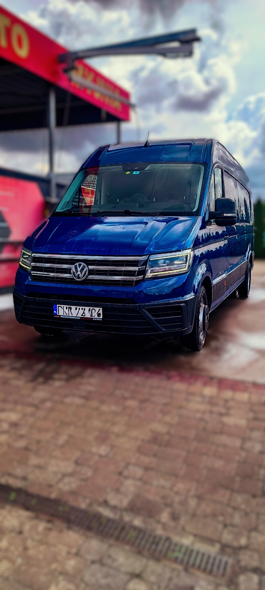 Niebieski bus Volkswagen Crafter po myciu, krople wody na lakierze, widok z przodu na tle myjni samochodowej.