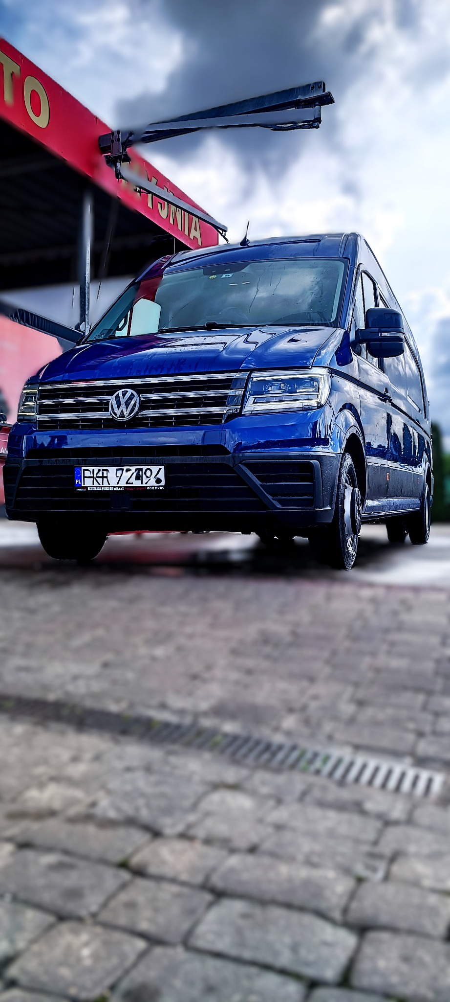 Niebieski bus Volkswagen Crafter po umyciu na myjni automatycznej, widoczne krople wody na karoserii i tablica rejestracyjna PKR 7Z496, na tle pochmurnego nieba.