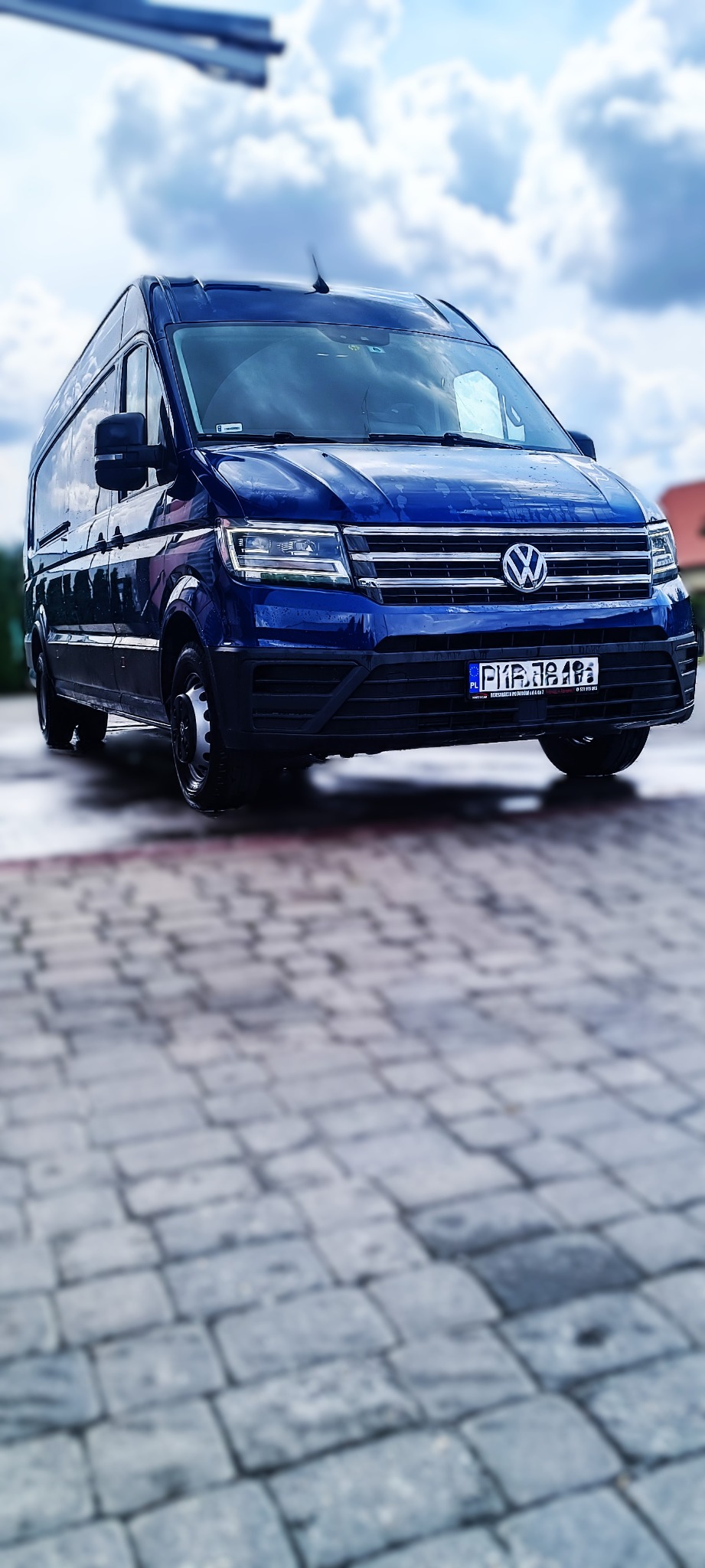 Niebieski bus Volkswagen Crafter zaparkowany na brukowanej nawierzchni, z widoczną tablicą rejestracyjną i odbiciami chmur na karoserii.