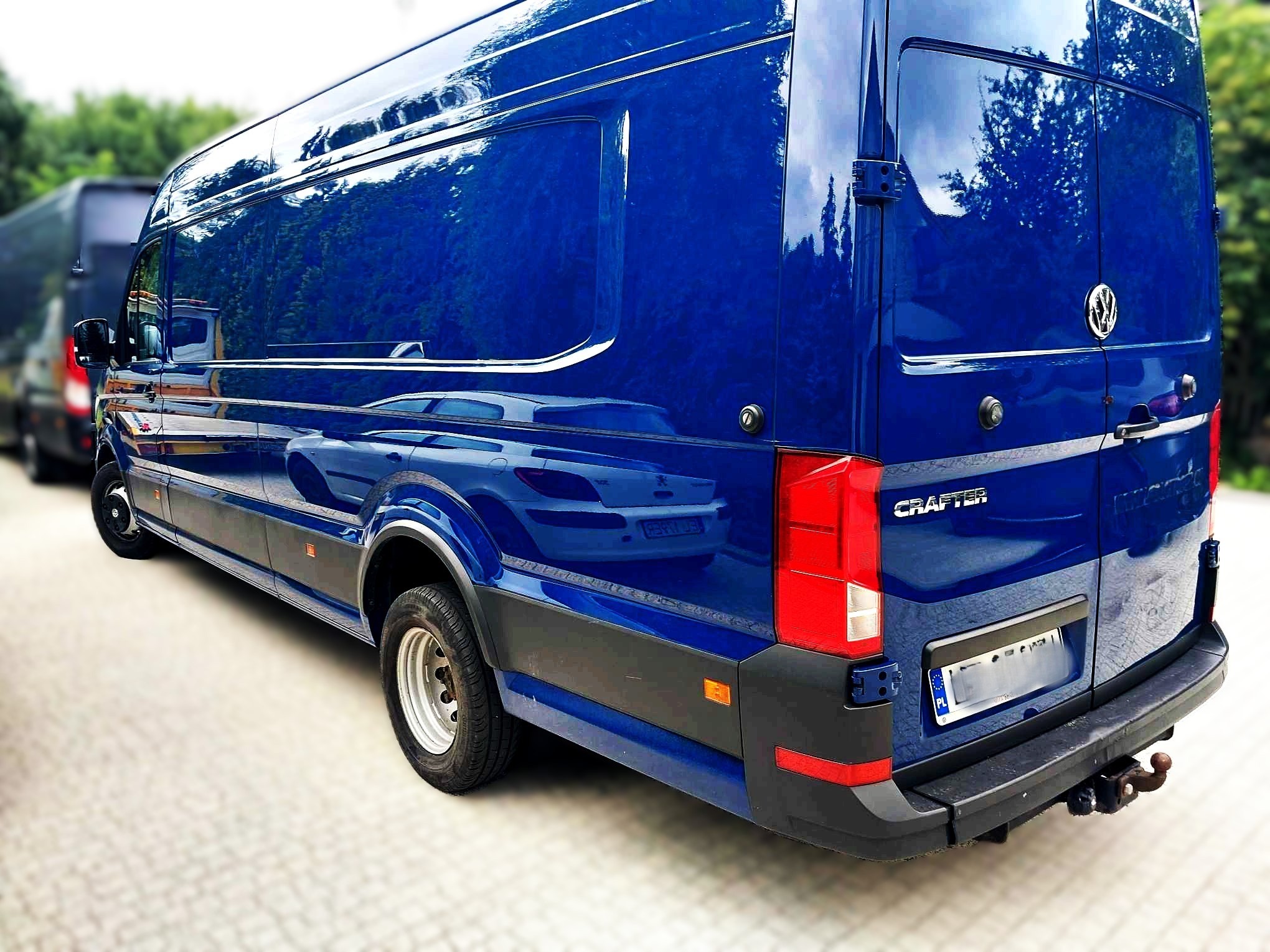 Niebieski bus Volkswagen Crafter z hakiem holowniczym, zaparkowany na jasnym bruku, z widocznym odbiciem otoczenia w lakierze.