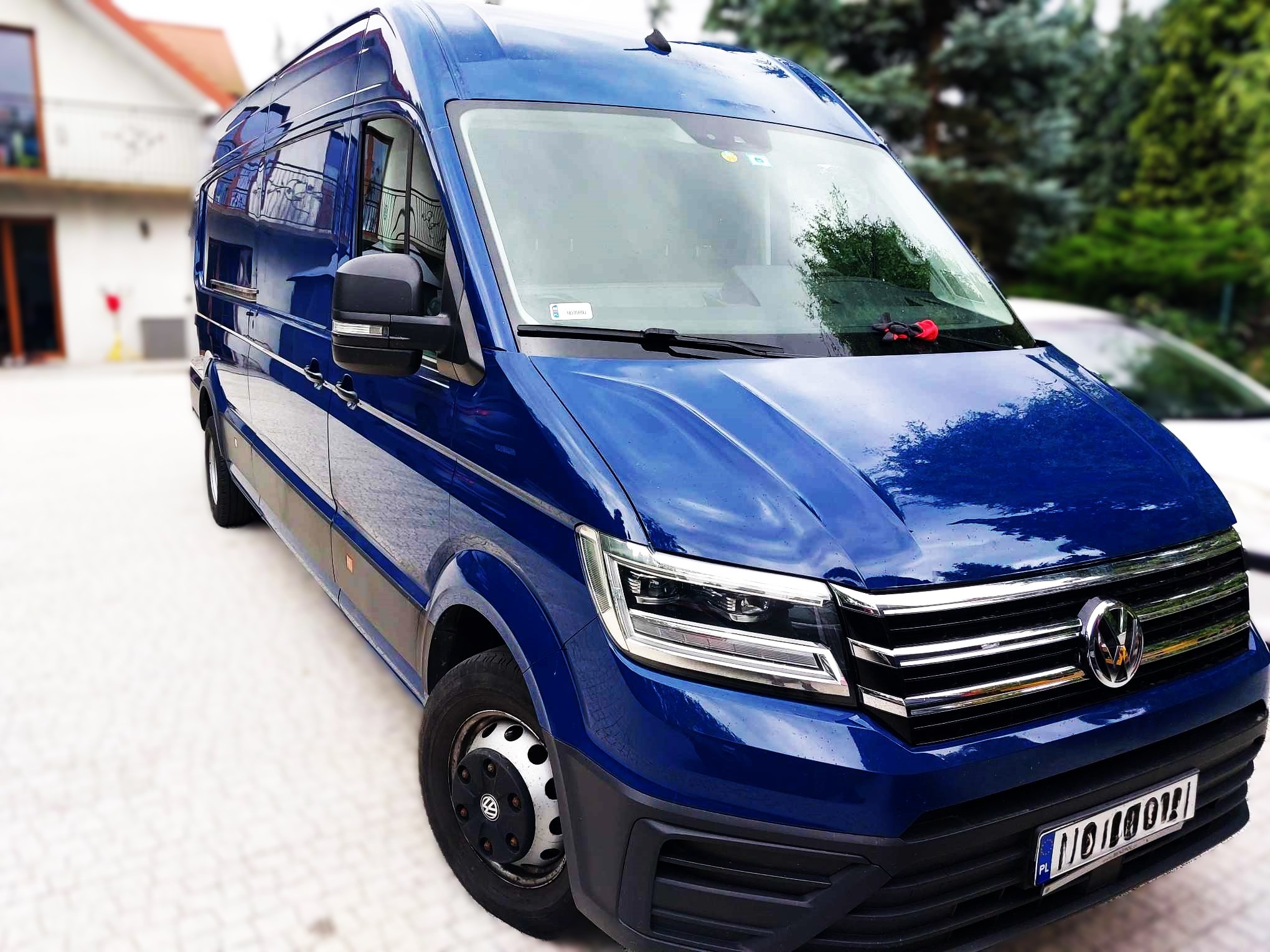 Niebieski Volkswagen Crafter z 2020 roku, widok z przodu na tle domu i drzew. Widoczne detale: chromowany grill, reflektory LED, tablica rejestracyjna i gumowa zabawka na desce rozdzielczej.
