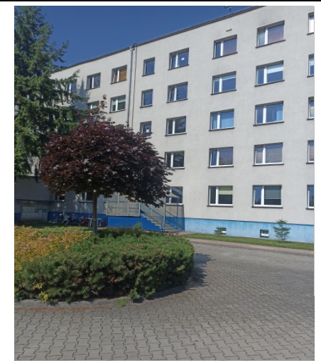 Szary budynek wielorodzinny z niebieskimi akcentami, widok z zewnątrz, z zadbanym terenem zielonym i drzewem o purpurowych liściach.