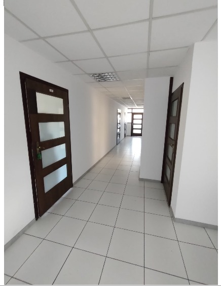 Długi korytarz biurowy z białymi ścianami, brązowymi drzwiami z przeszkleniami i podłogą wyłożoną jasnymi płytkami, widziany z perspektywy lekko skierowanej w dół.