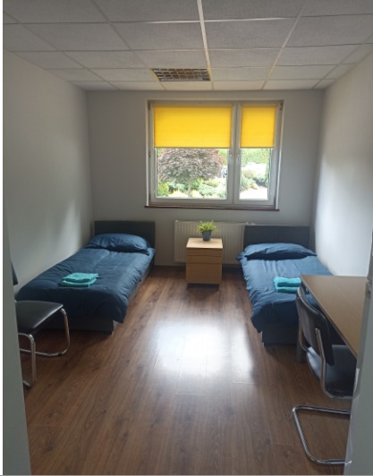 Pokój z dwoma łóżkami pojedynczymi z niebieską pościelą i turkusowymi ręcznikami, żółte rolety w oknie, drewniana podłoga i biurko z krzesłem.