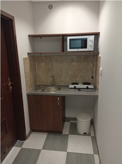 Aneks kuchenny z mikrofalówką, dwupalnikową kuchenką elektryczną i małym zlewem, widok z niskiego kąta, szaro-biała podłoga w kratkę.