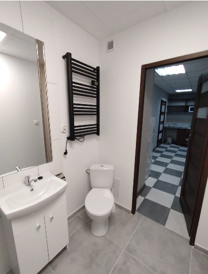 Jasna łazienka z umywalką na szafce, toaletą i czarnym grzejnikiem drabinkowym, widok na pomieszczenie z szachownicą na podłodze przez otwarte drzwi.