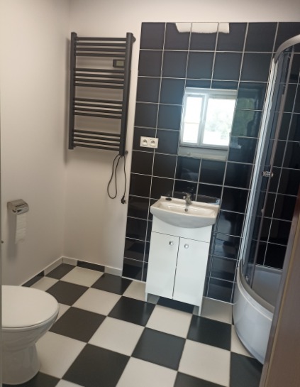 Wykończona łazienka z czarno-białą szachownicą na podłodze, białą szafką pod umywalką, kabiną prysznicową z czarnymi panelami i grzejnikiem drabinkowym na ścianie.