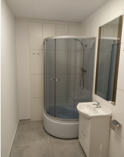 Wnętrze łazienki z kabiną prysznicową z zaokrąglonymi drzwiami, umywalką z szafką i lustrem w złotej ramie, całość utrzymana w bieli i szarościach.