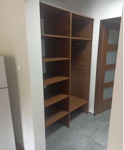 Wnękowa szafa z półkami i drążkiem na wieszaki, wykończona drewnem, widoczna z boku, z szarą podłogą i fragmentem białej lodówki po lewej stronie.