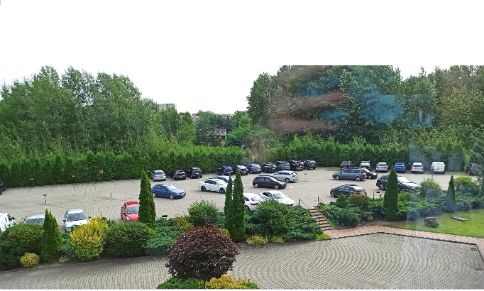Widok z góry na parking z wieloma samochodami, otoczony zielenią i drzewami, z widocznym budynkiem w tle.