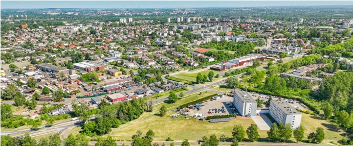 Panoramiczny widok z lotu ptaka na miasto z budynkami mieszkalnymi, biurowcami i zielonymi terenami, w tle widoczne drzewa i błękitne niebo.