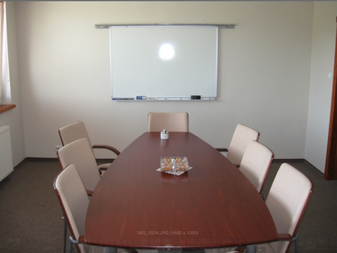 Sala konferencyjna z owalnym stołem z ciemnego drewna, krzesłami z jasną tapicerką, białą tablicą suchościeralną i drobnymi przekąskami na stole.