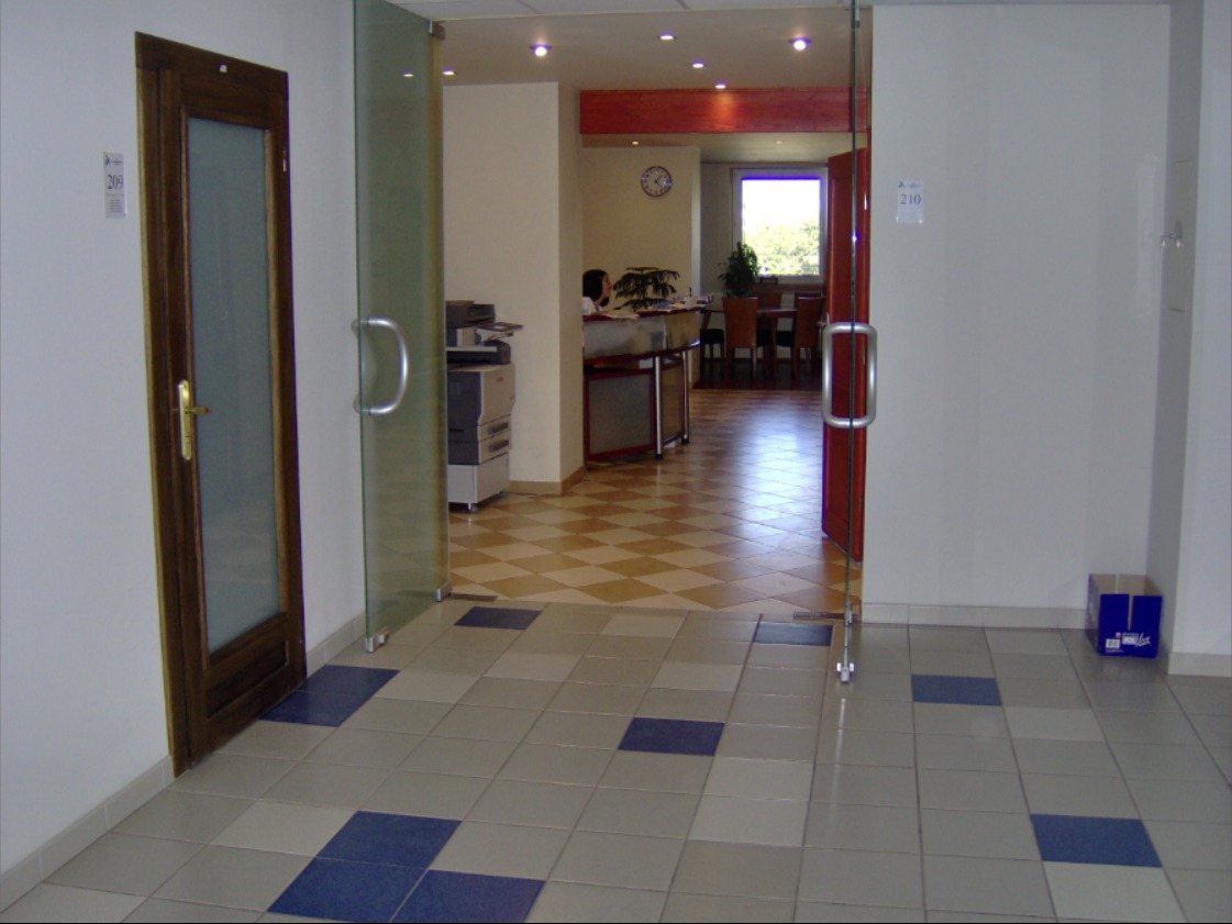 Korytarz biurowy z dwiema parami drzwi, jedna drewniana z szybą i druga szklana, widok na biuro z recepcją i salą konferencyjną, podłoga wyłożona płytkami w kolorach szarym i niebieskim.