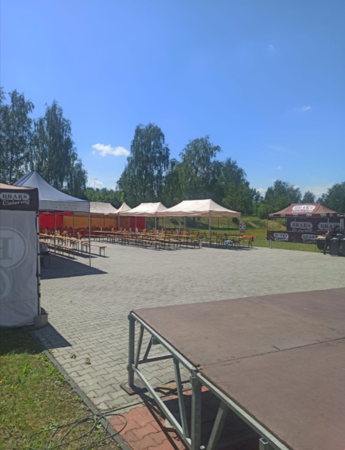 Przygotowane miejsce na imprezę plenerową z rzędami stołów pod białymi namiotami, widoczna scena i logo firmy cateringowej.
