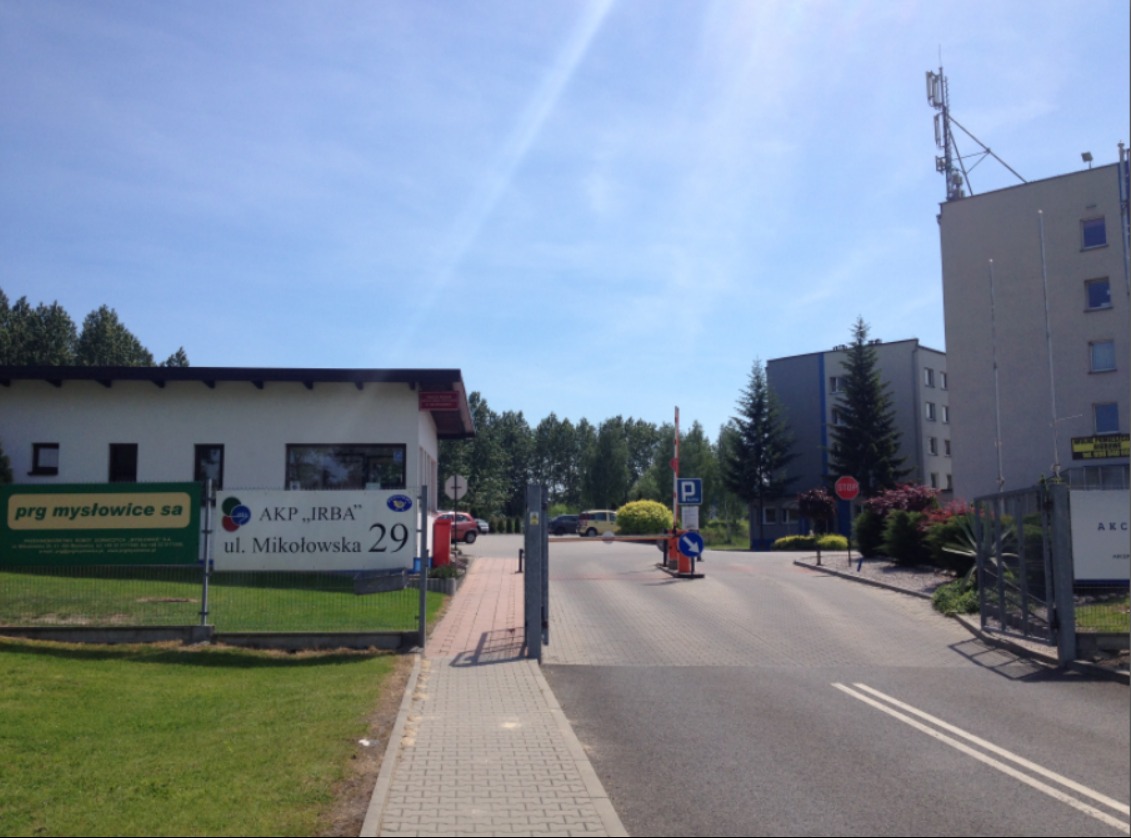 Widok na budynek z tablicą informacyjną 'AKP IRBA ul. Mikołowska 29', szlabanem i parkingiem w tle, w słoneczny dzień.