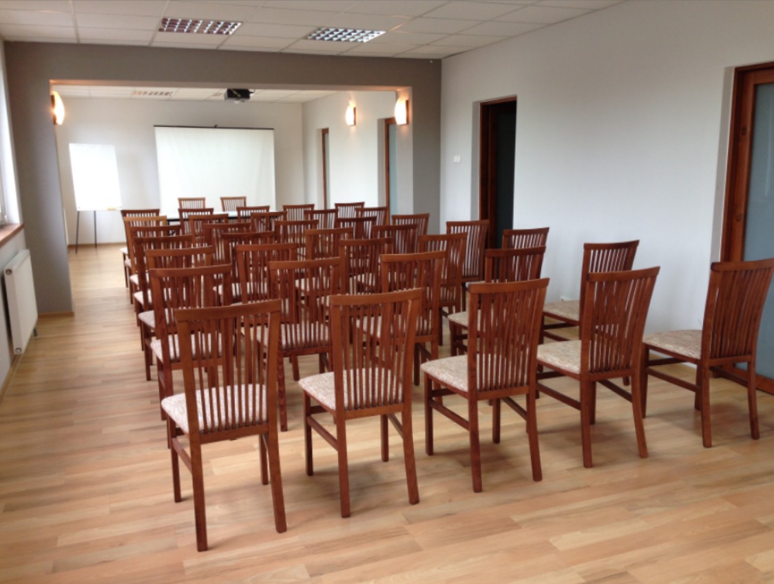 Sala konferencyjna z rzędami drewnianych krzeseł z beżowymi siedziskami, biały ekran projekcyjny w tle, widoczne lampy ścienne i jasna podłoga.