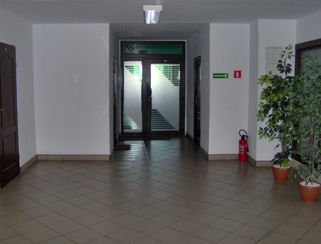 Jasny korytarz biurowca z roślinami w donicach, gaśnicą i przeszklonymi drzwiami z motywem schodów.