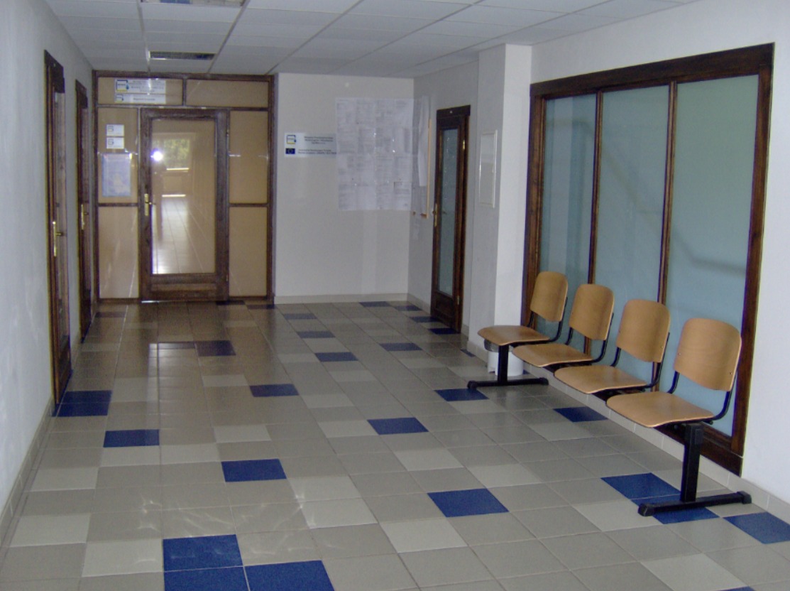 Poczekalnia z rzędem drewnianych krzeseł na metalowej podstawie, kafelki w szaro-niebieską szachownicę na podłodze, szklane drzwi i tablica informacyjna na ścianie.