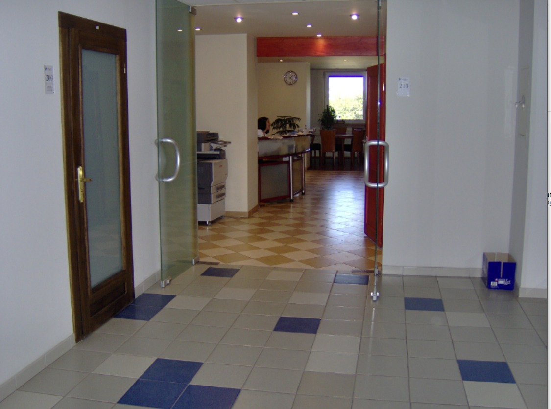 Wnętrze biura z widocznymi drzwiami z brązową ramą i przeszklonymi drzwiami, w tle recepcja z pracownikiem i salą konferencyjną.