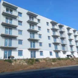 Wielopiętrowy budynek z szarą elewacją i licznymi balkonami, okna zabezpieczone folią ochronną, teren wokół w trakcie prac ziemnych, widoczne materiały budowlane.