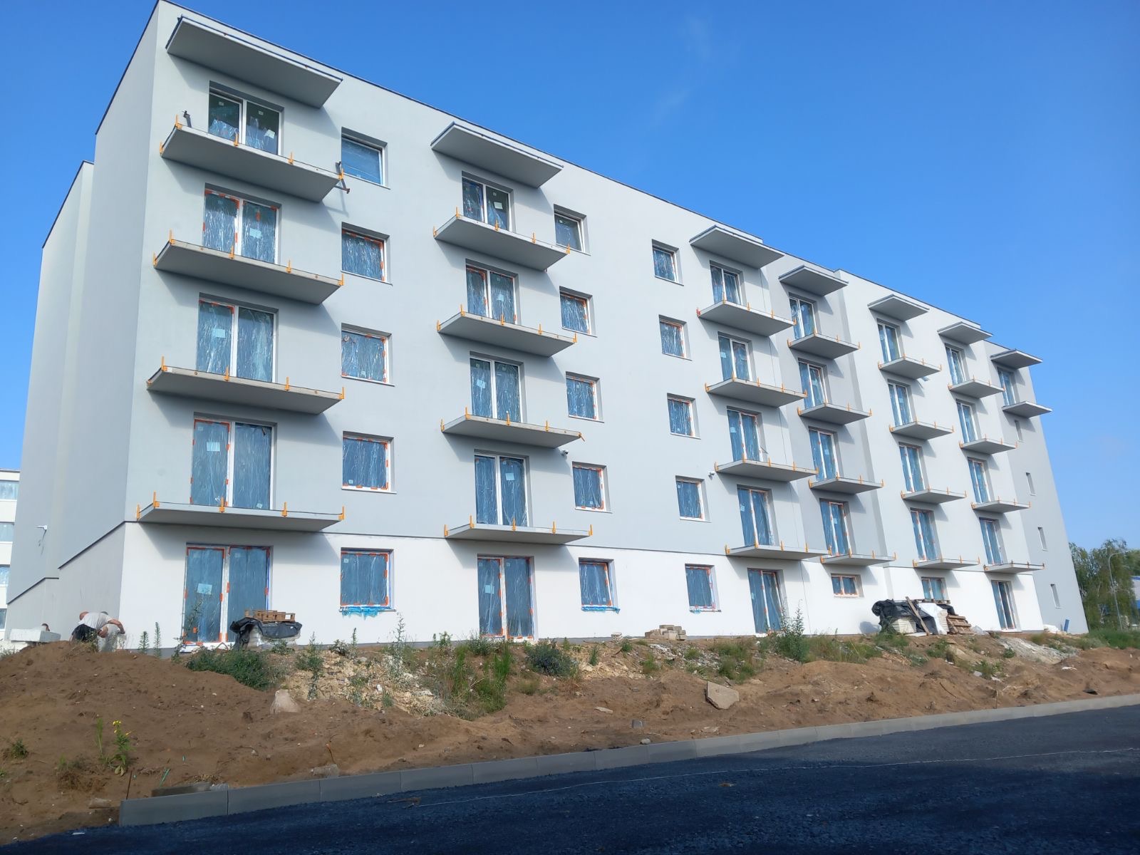 Wielopiętrowy budynek z szarą elewacją i licznymi balkonami, okna zabezpieczone folią ochronną, teren wokół w trakcie prac ziemnych, widoczne materiały budowlane.