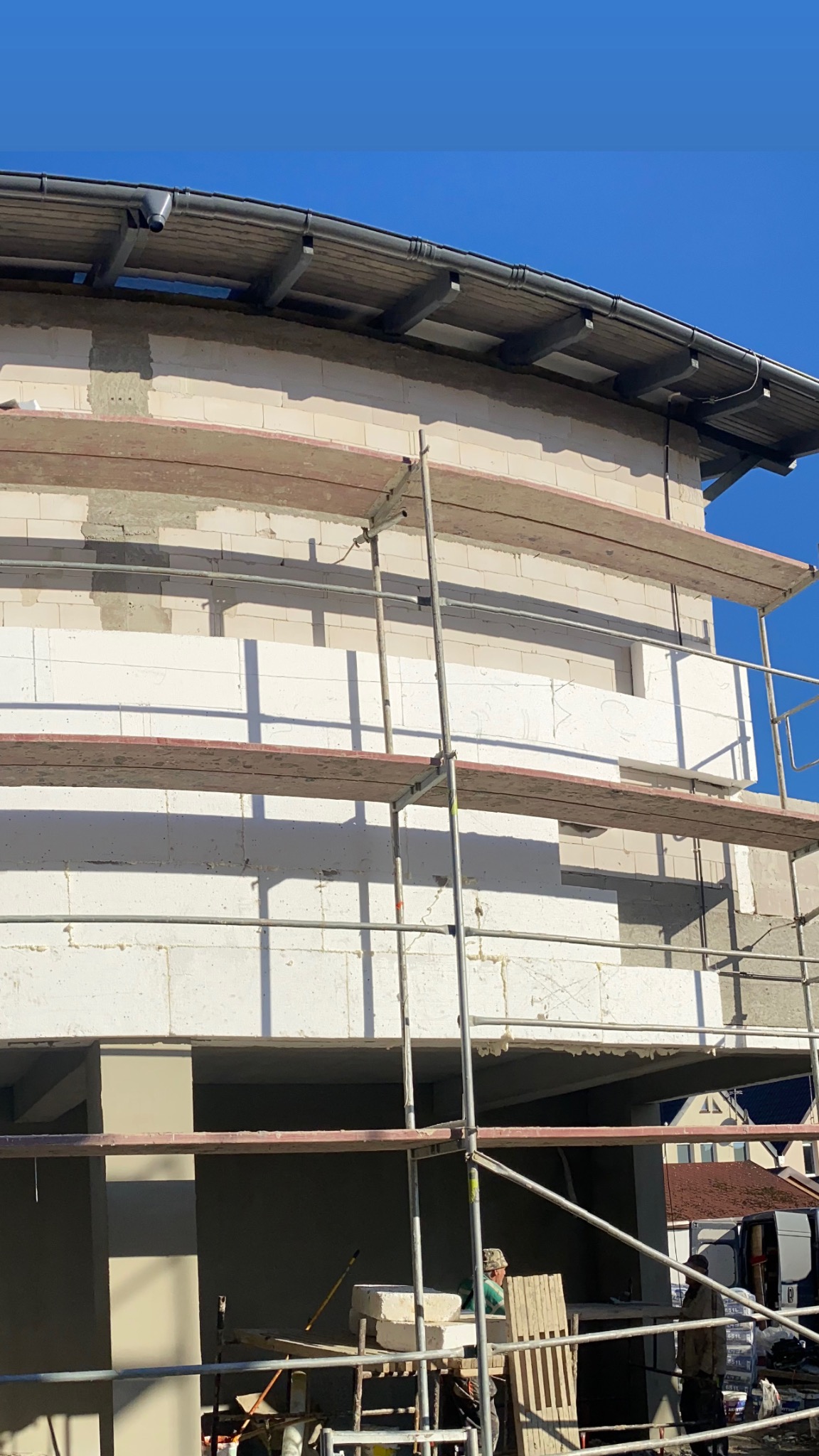 Budynek w trakcie ocieplania, widoczne rusztowania, białe bloki izolacyjne, częściowo otynkowana ściana i elementy dachu na tle błękitnego nieba.