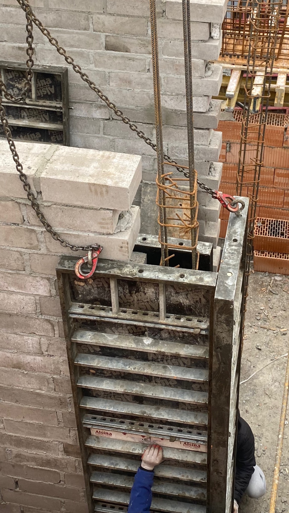 Wznoszenie ściany z bloczków betonowych z użyciem szalunku systemowego Alisina i zbrojenia stalowego, widoczne łańcuchy i haki mocujące, w tle mur z cegły dziurawki.