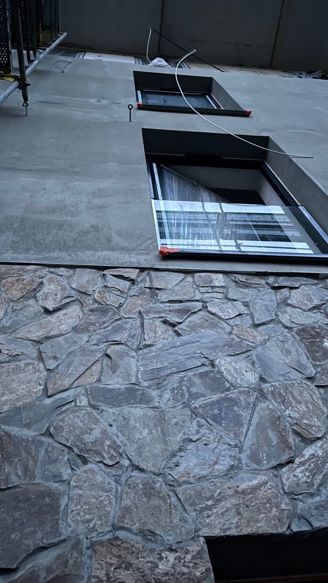 Fragment elewacji budynku z oknami zabezpieczonymi folią, z wykończeniem dolnej części kamieniem elewacyjnym i świeżo położonym tynkiem na górze, widoczne rusztowanie.