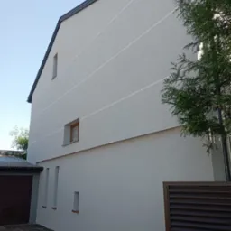 Jasnoszara elewacja domu jednorodzinnego z widocznymi poziomymi pasami dekoracyjnymi, małymi oknami i brązową bramą garażową po lewej stronie, częściowo zasłonięta drzewem po prawej.