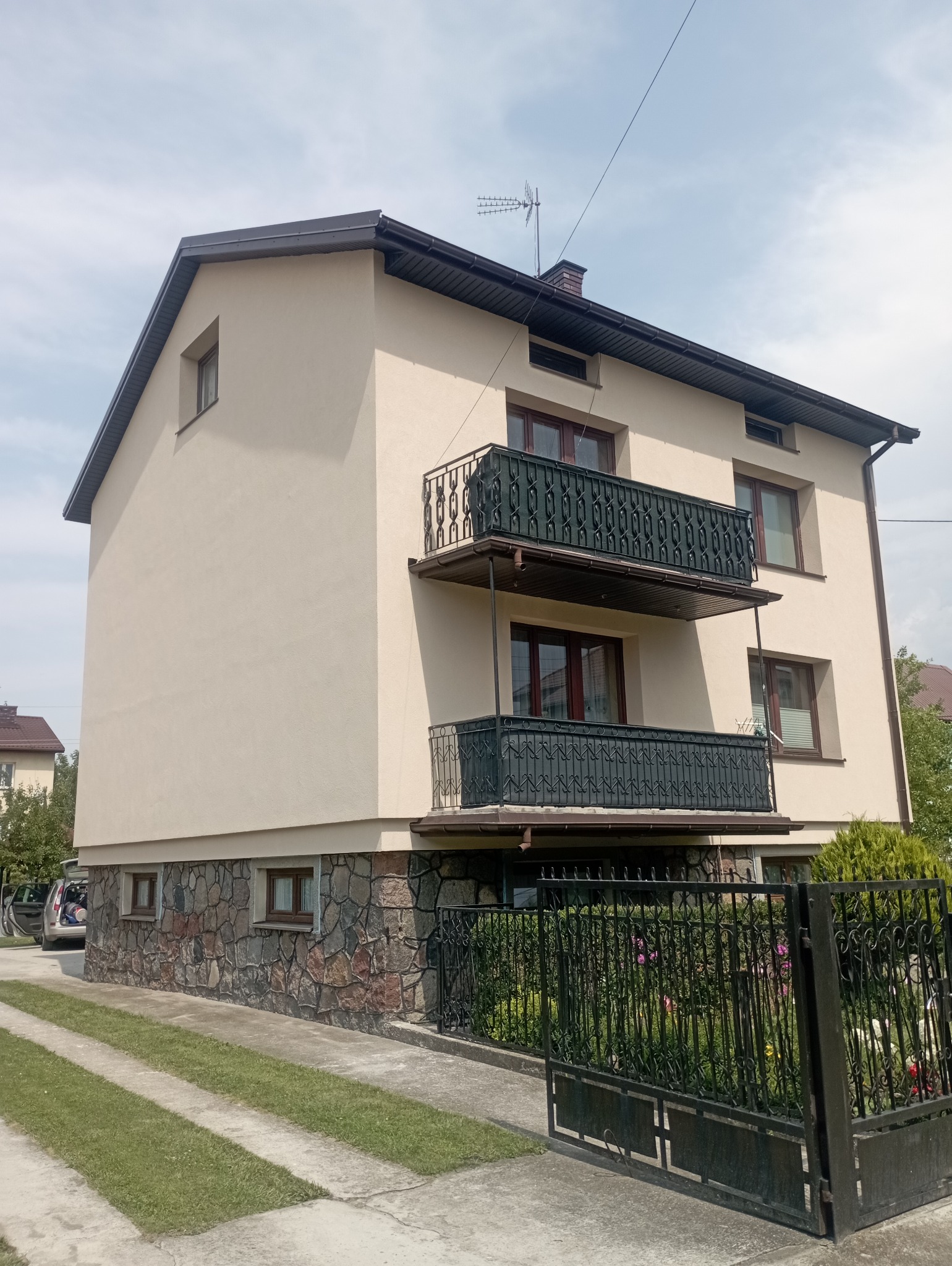 Dwupiętrowy dom z beżową elewacją, dwoma kutymi balkonami i kamienną podmurówką, widziany z poziomu ulicy w słoneczny dzień.