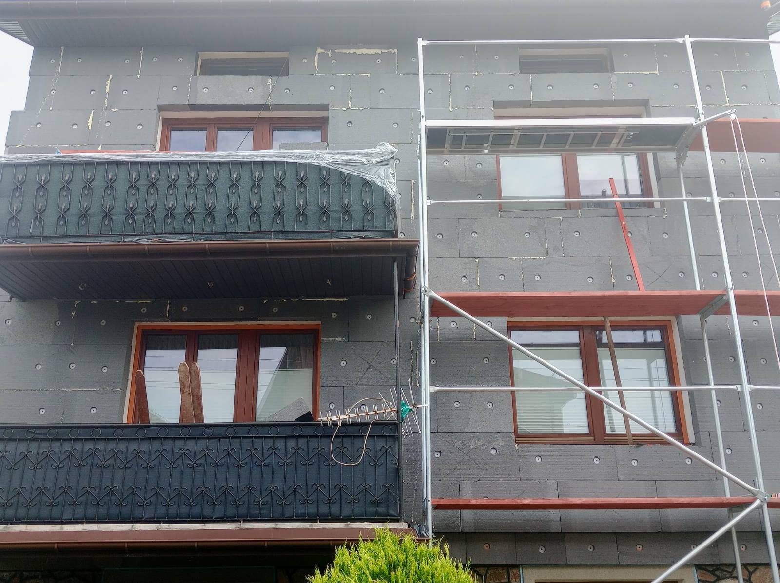 Dom w trakcie ocieplania szarym styropianem, widoczne rusztowanie i balkon z czarną balustradą, fragment anteny na balkonie.