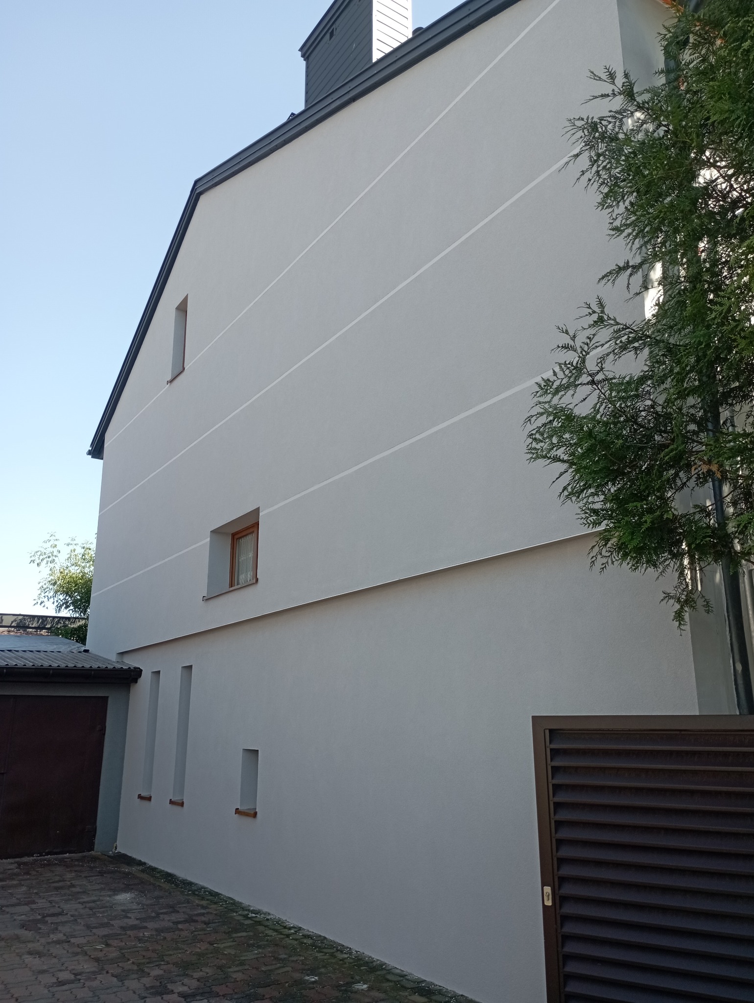 Jasnoszara elewacja domu jednorodzinnego z widocznymi poziomymi pasami dekoracyjnymi, małymi oknami i brązową bramą garażową po lewej stronie, częściowo zasłonięta drzewem po prawej.