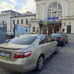 Dwa samochody taxi stojące przed budynkiem dworca kolejowego w Tarnowie, widoczne detale takie jak taksometr na dachu, tablice rejestracyjne i naklejki informacyjne na karoserii.