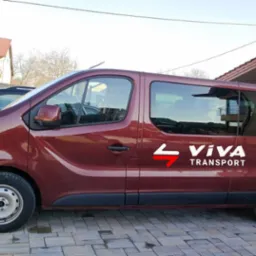 Bordowy bus z logo firmy transportowej Viva Transport zaparkowany na brukowanym podjeździe przed budynkiem.
