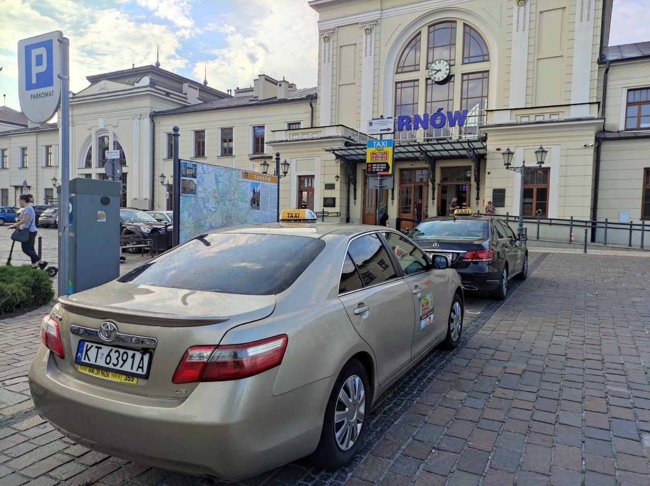 Dwa samochody taxi stojące przed budynkiem dworca kolejowego w Tarnowie, widoczne detale takie jak taksometr na dachu, tablice rejestracyjne i naklejki informacyjne na karoserii.