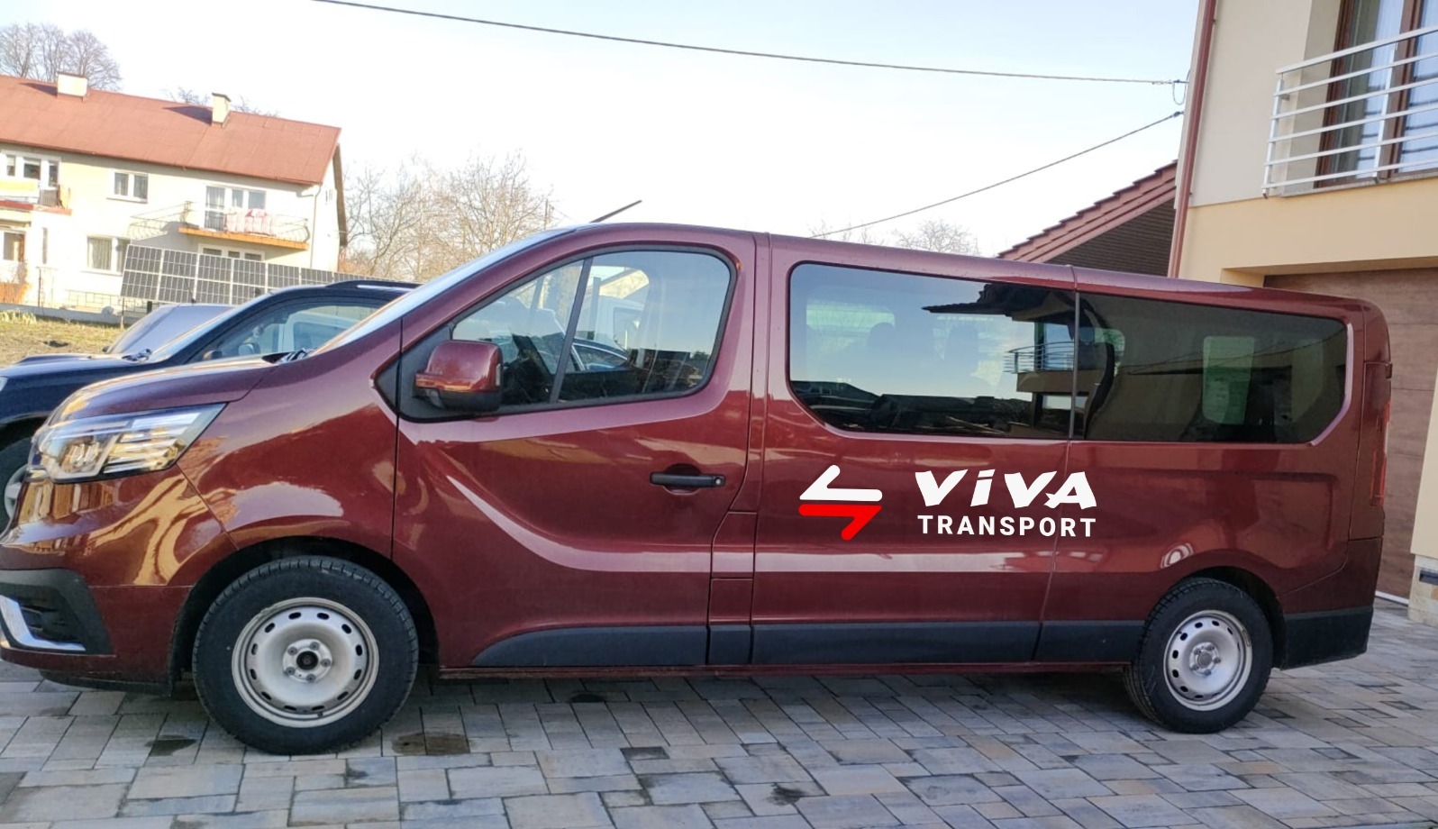 Bordowy bus z logo firmy transportowej Viva Transport zaparkowany na brukowanym podjeździe przed budynkiem.