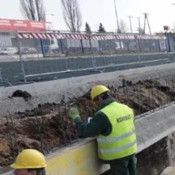 Dwóch pracowników w żółtych kamizelkach i kaskach, przy betonowym wykopie z wystającymi prętami zbrojeniowymi, zabezpieczonym barierkami.
