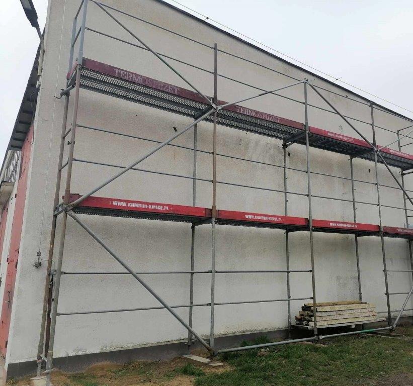 Budynek w trakcie ocieplania, widoczne rusztowanie z czerwonymi elementami oraz stos płyt izolacyjnych u podstawy.