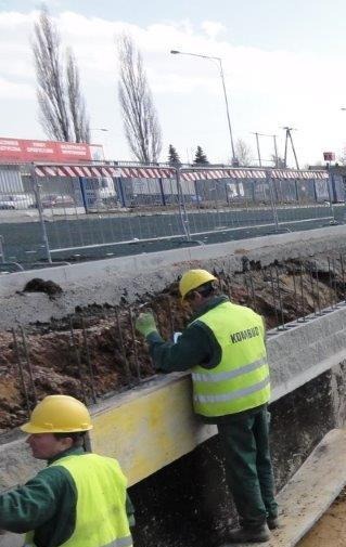 Dwóch pracowników w żółtych kamizelkach i kaskach, przy betonowym wykopie z wystającymi prętami zbrojeniowymi, zabezpieczonym barierkami.