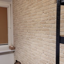 Perfekcyjnie Profesionalnie - Ściana wykończona tapetą imitującą cegłę, widoczny fragment okna z roletą, metalowy regał i osoba w czarnej koszulce, prawdopodobnie podczas pracy.