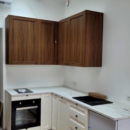 Perfekcyjnie Profesionalnie - Nowoczesna kuchnia w odcieniach bieli i drewna z marmurowym blatem, widok na zabudowę kuchenną z drewnianymi szafkami górnymi i białymi dolnymi, drewniana belka z oświetleniem na suficie.