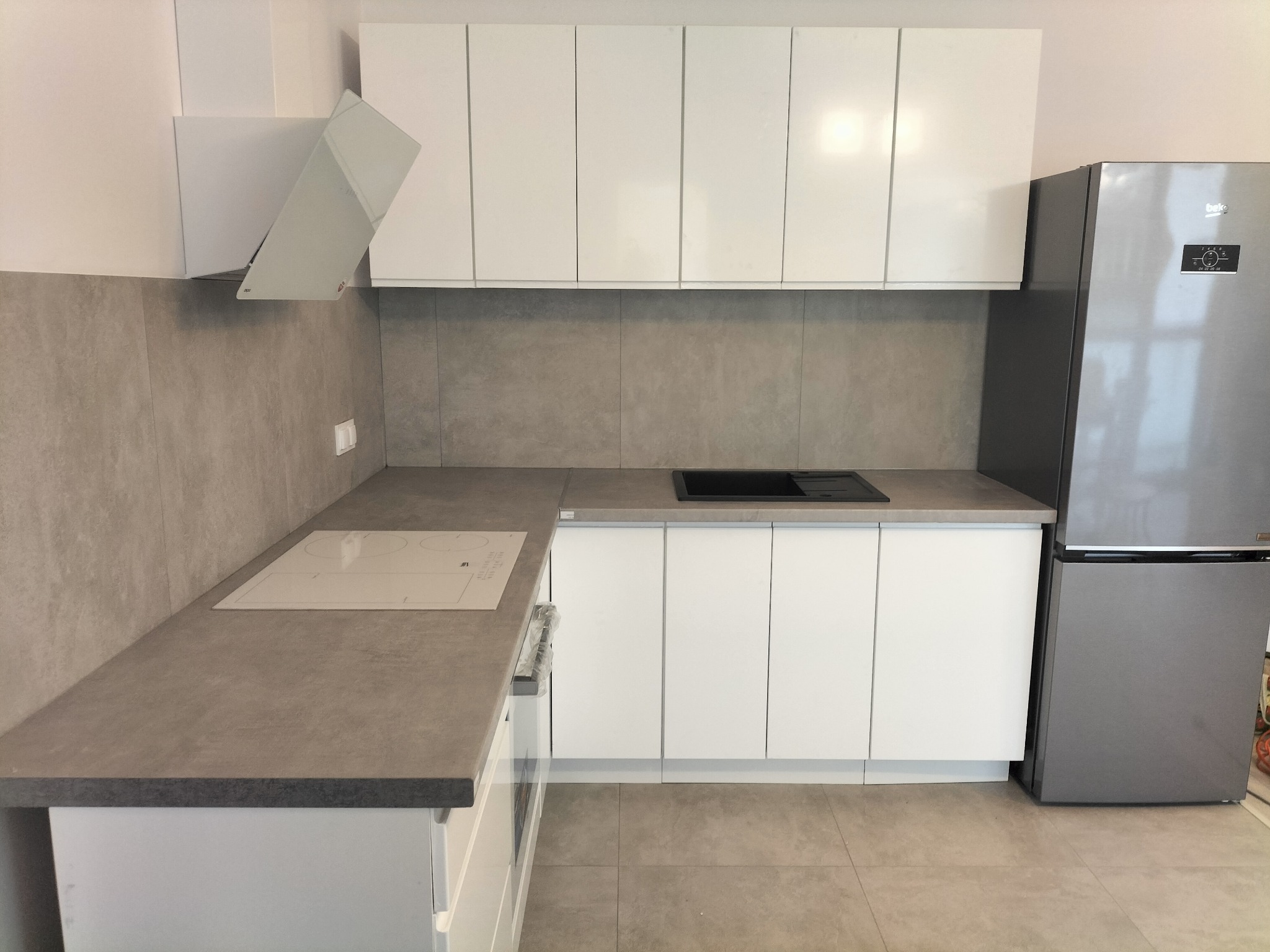 Nowoczesna, jasna kuchnia z białymi szafkami i szarym blatem, widok na płytę indukcyjną, okap i lodówkę Beko