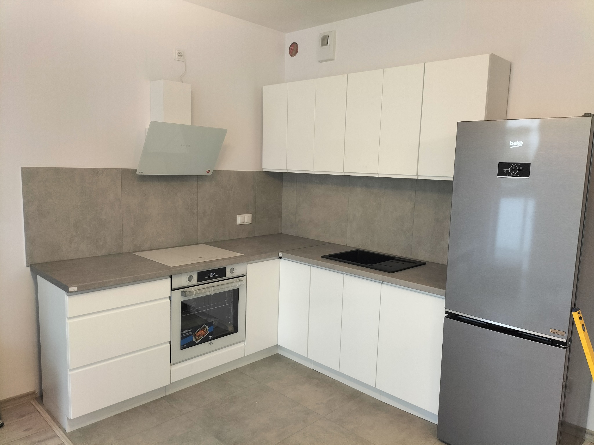 Nowoczesna, jasna kuchnia z białymi szafkami, szarym blatem i srebrną lodówką Beko. Na ścianie szare płytki. Widoczny poziomica przy lodówce.