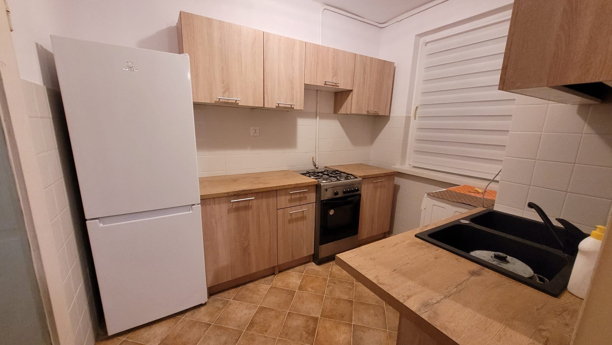 Kuchnia po remoncie: zabudowa meblowa z drewnopodobnymi frontami, biała lodówka, ciemny zlew, brązowa podłoga z płytek, białe rolety w oknie.