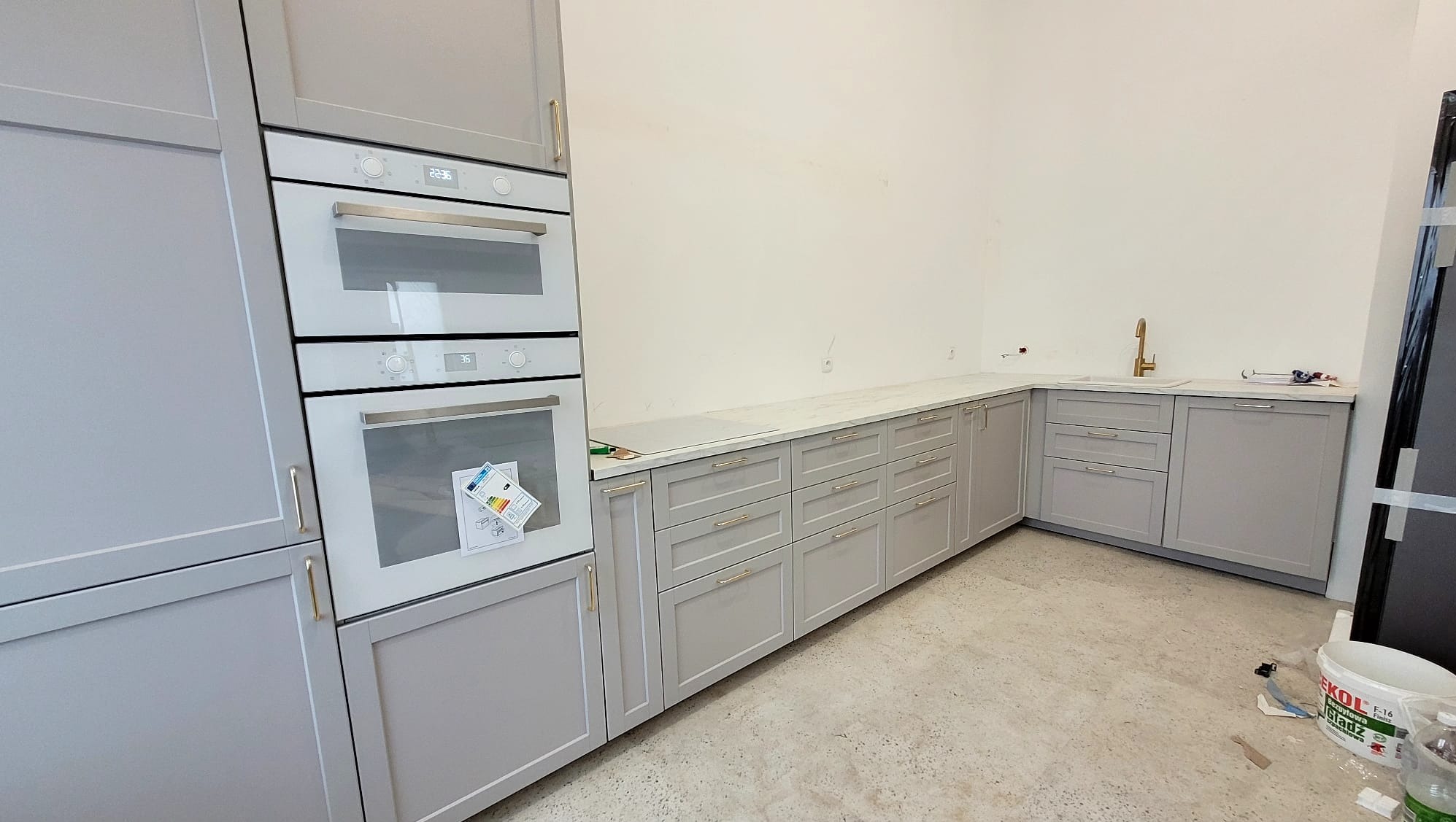 Szare, minimalistyczne meble kuchenne z białym blatem i złotą armaturą, widoczne naklejki na piekarniku, surowy beton na podłodze.
