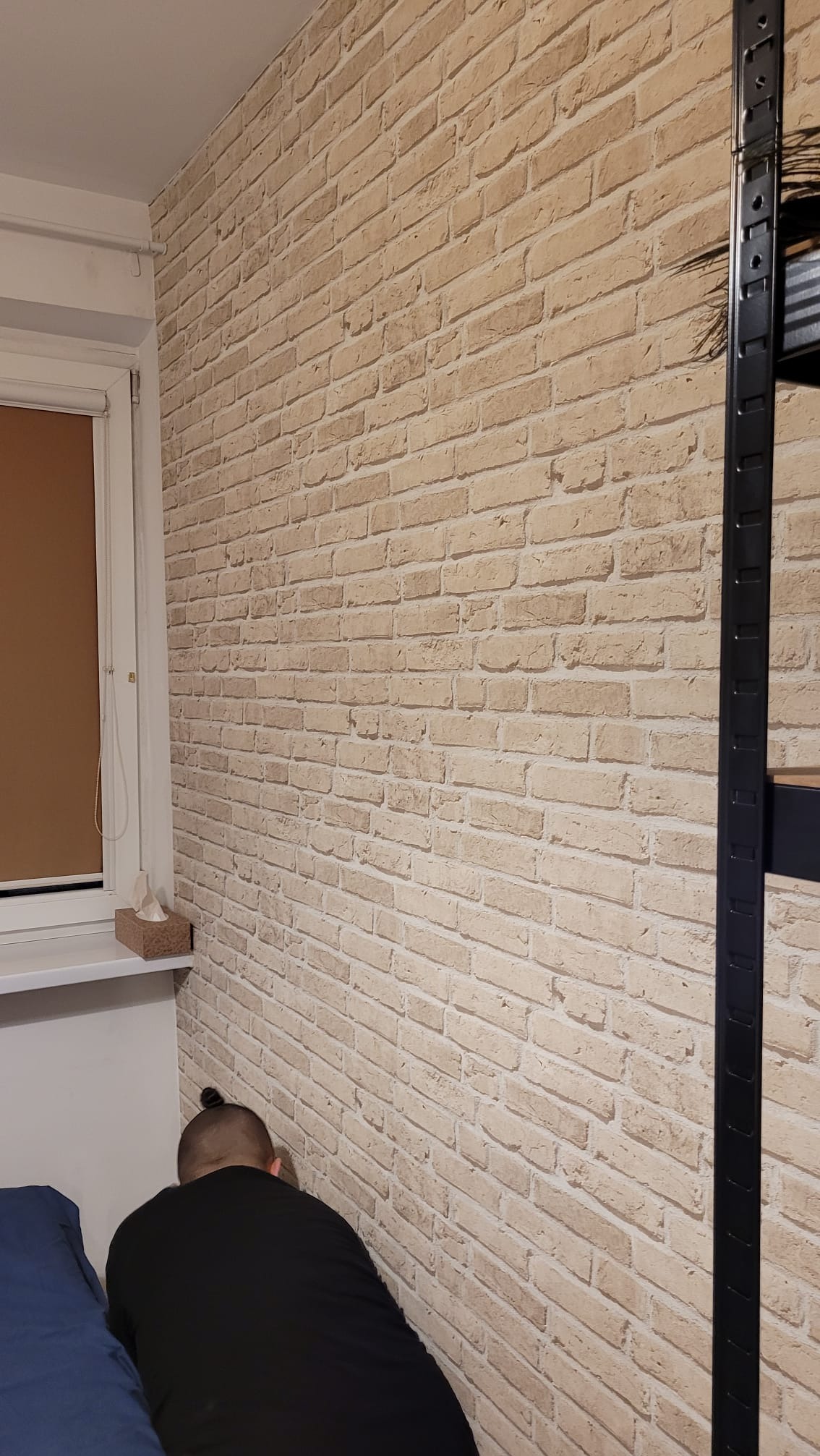Ściana wykończona tapetą imitującą cegłę, widoczny fragment okna z roletą, metalowy regał i osoba w czarnej koszulce, prawdopodobnie podczas pracy.
