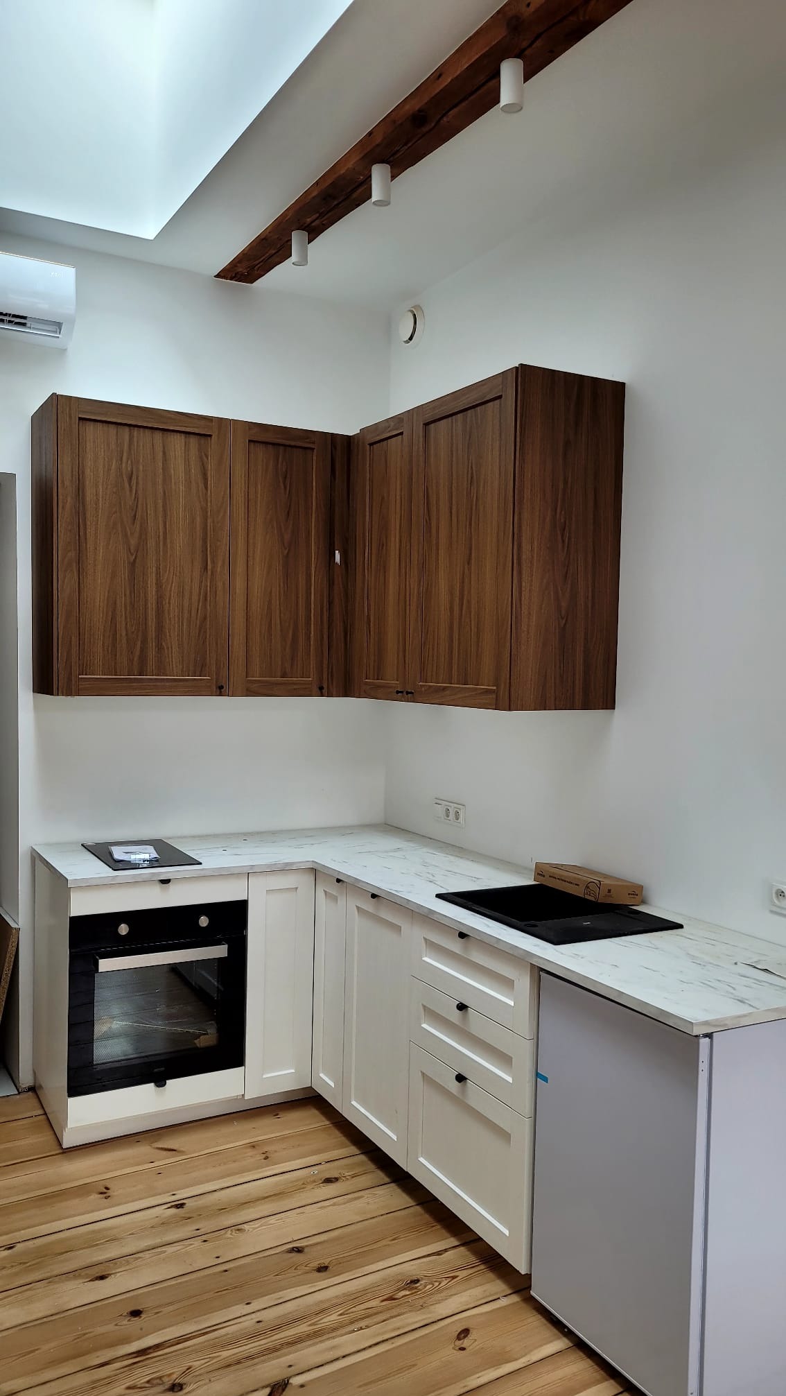 Nowoczesna kuchnia w odcieniach bieli i drewna z marmurowym blatem, widok na zabudowę kuchenną z drewnianymi szafkami górnymi i białymi dolnymi, drewniana belka z oświetleniem na suficie.