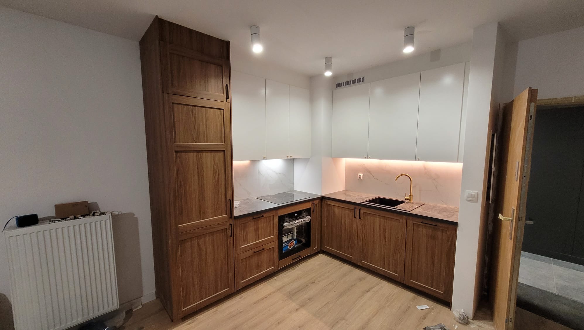 Nowoczesna kuchnia z białymi górnymi szafkami i dolnymi szafkami z drewna, czarnym zlewem i złotą baterią, oświetleniem LED pod szafkami i trzema punktowymi lampami sufitowymi.