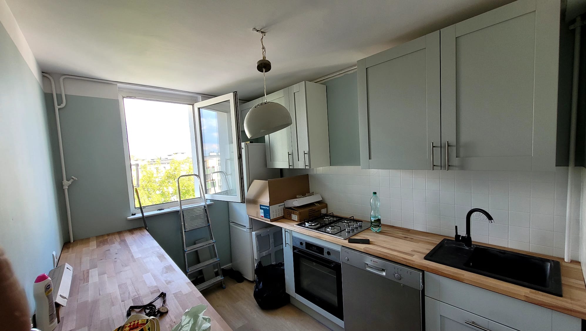 Kuchnia w trakcie remontu: szare szafki kuchenne, drewniany blat, białe płytki, okno z widokiem na zieleń, drabina oparta o ścianę, kartony i butelka na blacie, wisząca lampa, czarny zlew i srebrne...